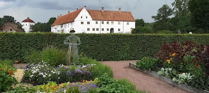 En slottsträdgård med färgglada odlingar och en staty i hönsnät. I bakgrunden syns ett vitt slott.