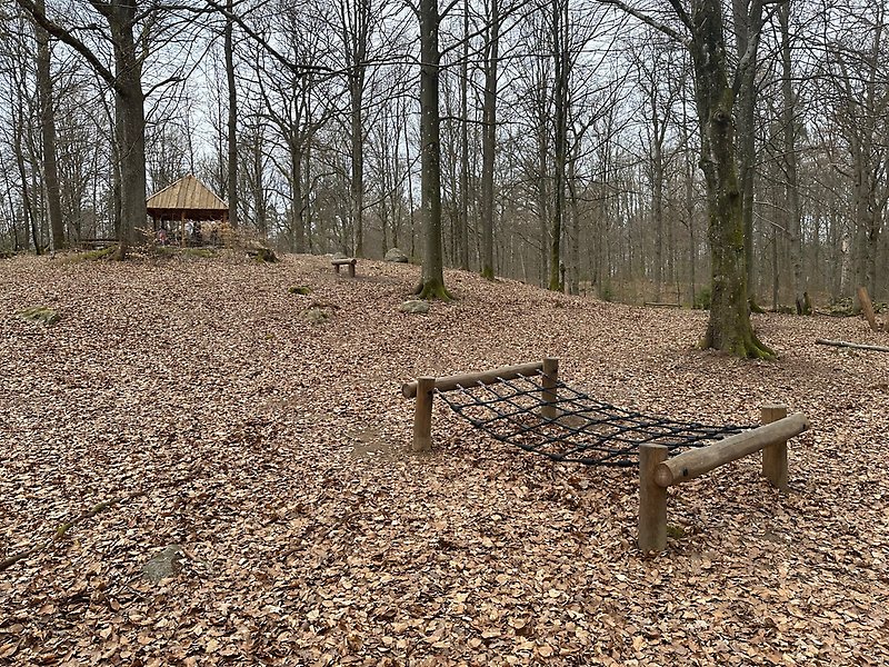 Lekplats med hinder i skogen och en grillplats med tak i bakgrunden.